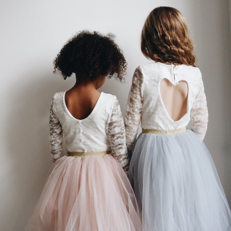 Op de afbeelding: Twee meisjes dragen witte kant tops met lange mouwen en tule rokken. De ene rok is roze en de andere is blauw. De witte top heeft een hartvormige uitsnijding in de rug.