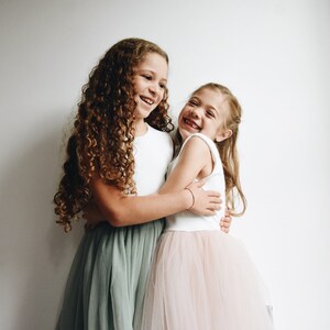 May include: Two young girls, one with long curly brown hair wearing a white top and a sage green tulle skirt, and the other with long straight blonde hair wearing a white top and a blush pink tulle skirt, are hugging each other and smiling.
