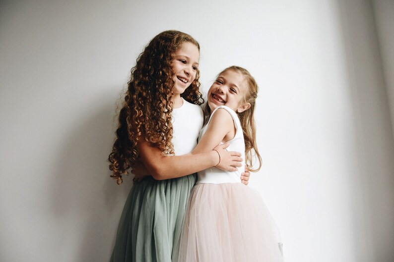 May include: Two young girls, one with long curly brown hair wearing a white top and a sage green tulle skirt, and the other with long straight blonde hair wearing a white top and a blush pink tulle skirt, are hugging each other and smiling.