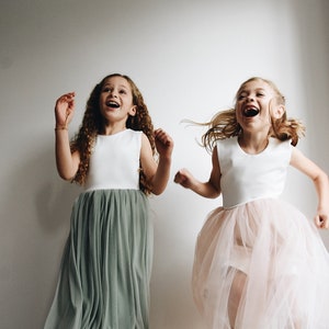 May include: Two girls wearing white sleeveless tops with tulle skirts. One girl is wearing a sage green skirt and the other is wearing a pink skirt. Both girls are smiling and jumping in the air.