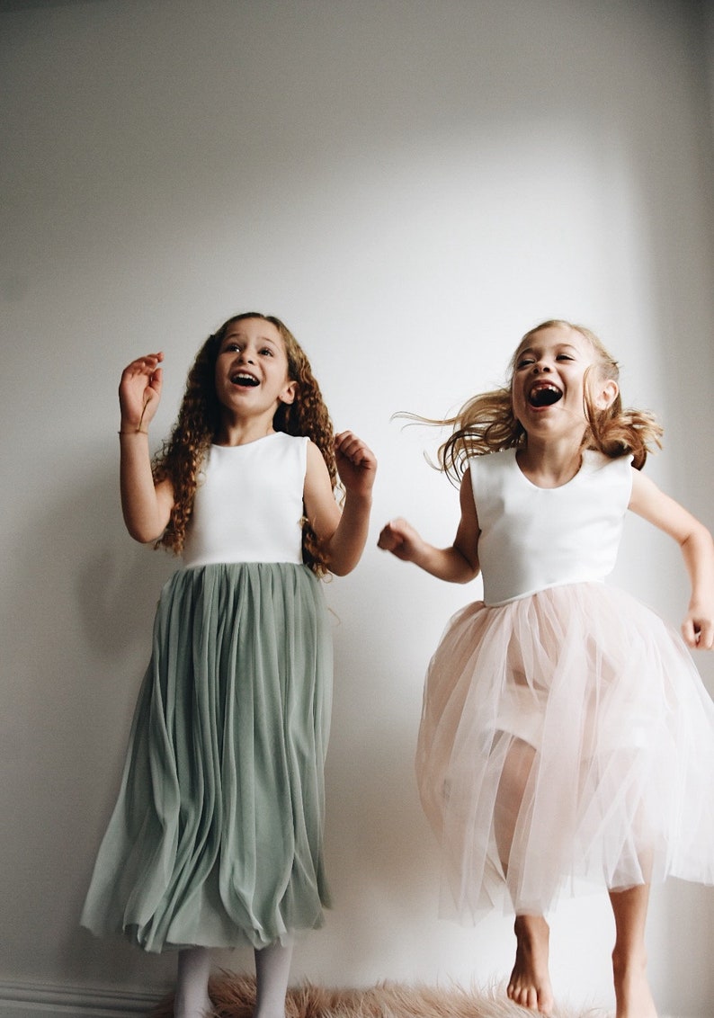 May include: Two girls wearing white sleeveless tops with tulle skirts. One girl is wearing a sage green skirt and the other is wearing a pink skirt. Both girls are smiling and jumping in the air.