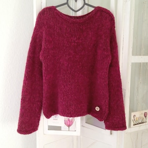 May include: A deep red knitted sweater with a crew neck and long sleeves. The textured jumper features a small, round button detail on the lower left. The garment is suspended from a heart-shaped hanger.