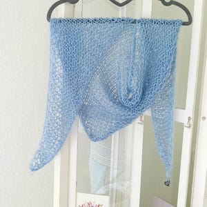 Puede incluir: Un chal azul claro de tejido abierto se exhibe en una percha. El chal tiene forma triangular con una textura delicada y aireada. El color es un azul pastel suave y la tela parece ligera y fluida. Un pequeño dije en forma de corazón cuelga de una de las puntas.