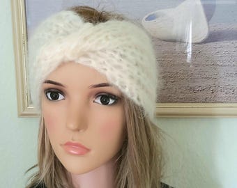 Op de afbeelding: Crèmekleurige, gebreide hoofdband met een gedraaid knoopontwerp. De hoofdband is gemaakt van een zacht, pluizig garen. De afbeelding toont de hoofdband op een mannequinhoofd met blond haar, tegen een achtergrond van een ingelijst kunstwerk.
