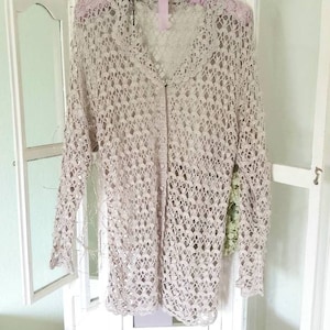Könnte beinhalten: Ein beigefarbener Häkelcardigan mit Knopfleiste und langen Ärmeln. Der Cardigan hängt an einem weißen Kleiderbügel vor einem weißen Hintergrund.
