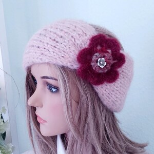 Puede incluir: Boina de punto rosa claro con un adorno floral rojo oscuro. La flor tiene un botón plateado en el centro. La boina está hecha de un hilo suave y texturizado, ideal para el clima frío. Un accesorio elegante y cálido.