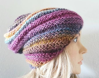 May include: A hand-knitted beanie hat in a mix of colours including purple, blue, yellow, and brown. The hat is displayed on a mannequin head with blonde hair, highlighting the textured knit design. The hat is a stylish accessory.