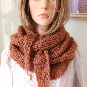 Puede incluir: Bufanda marrón tejida con un lazo y una etiqueta de madera. La bufanda está hecha de un material de punto grueso y está drapeada alrededor del cuello de un maniquí. El fondo es de color claro.