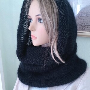 May include: A black knitted hood and cowl combination. The hood is designed to frame the face, while the cowl provides warmth around the neck. The knitted texture is visible, suggesting a handmade or artisanal item. The hood is displayed on a mannequin.