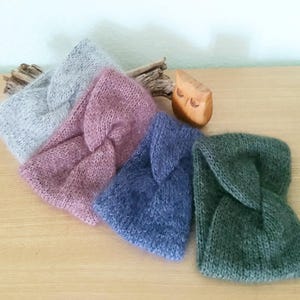 Könnte beinhalten: Vier gestrickte Stirnbänder in Grau-, Rosa-, Blau- und Grüntönen liegen auf einer Holzoberfläche. Jedes Stirnband hat ein verdrehtes Knotendesign. Eine kleine Holz-Eulenfigur und Treibholz sind ebenfalls vorhanden.