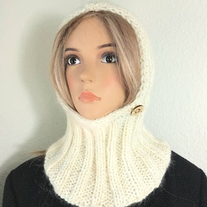 May include: Cream-colored knitted hood with a ribbed cowl neck and a wooden button closure. The hood is made of a soft, textured yarn and is designed to keep the head and neck warm. The hood is displayed on a mannequin.