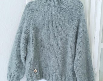 May include: A light gray, hand-knitted sweater with a high, ribbed collar and long sleeves. The sweater has a relaxed fit and a small, gold-colored button detail near the hem. The garment is displayed on a black hanger against a white background.