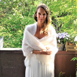 May include: A white knitted shawl with a delicate lace pattern and a fringe border. The shawl is draped over a person wearing a white dress.