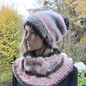 Könnte beinhalten: Handgestricktes Mützen- und Schal-Set. Die Mütze ist in Rosa-, Grau- und Blautönen gestreift, mit einem dunklen Bommel. Der Schal hat ein ähnliches Farbschema mit braunen Akzenten. Das Set wird auf einer Schaufensterpuppe präsentiert.