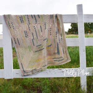 Könnte beinhalten: Eine beige Häkeldecke mit geometrischem Muster, drapiert über einem weißen Zaun. Die Decke hat Akzente in Rosa, Gelb, Grün und Blau. Die Decke besteht aus einem weichen Material mit offenem Gewebe.