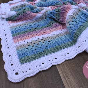 May include: A crocheted baby blanket with a white border and stripes of pink, blue, green, and white. The blanket is folded and lying on a wooden surface. The text "a Dolly Plum pattern" is visible in the lower right corner of the image.