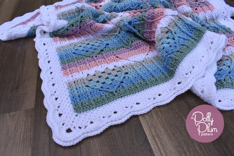 May include: A crocheted baby blanket with a white border and stripes of pink, blue, green, and white. The blanket is folded and lying on a wooden surface. The text "a Dolly Plum pattern" is visible in the lower right corner of the image.
