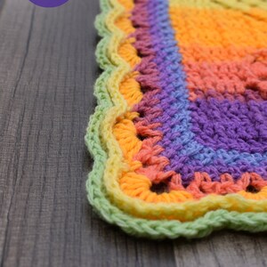 May include: A close-up of a colorful crocheted blanket with a scalloped edge. The blanket is made in a rainbow pattern with shades of green, yellow, orange, pink, and purple. The blanket is lying on a wooden surface.