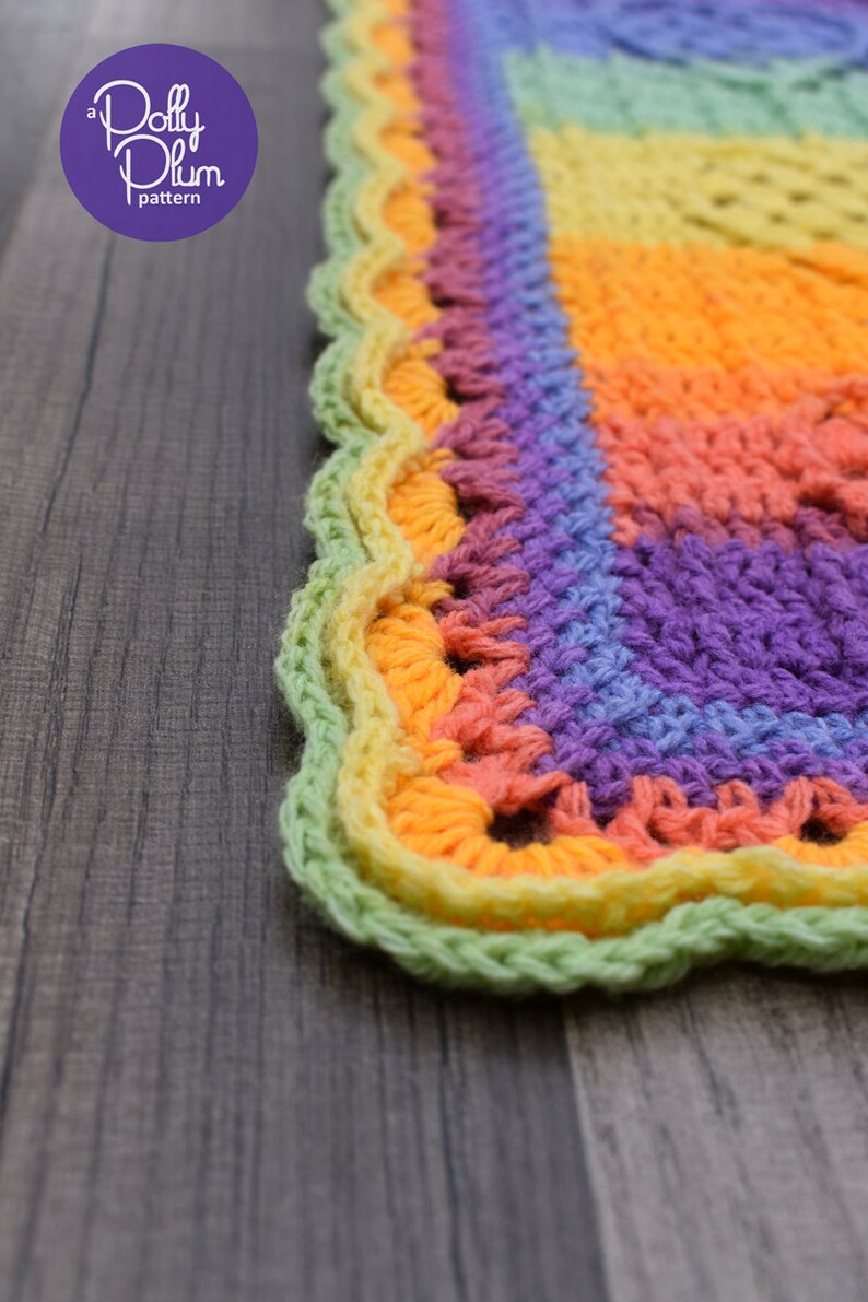 May include: A close-up of a colorful crocheted blanket with a scalloped edge. The blanket is made in a rainbow pattern with shades of green, yellow, orange, pink, and purple. The blanket is lying on a wooden surface.