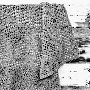May include: A gray crocheted blanket with a geometric pattern is draped over a weathered white fence. The blanket's texture is visible, with open spaces creating a delicate design. The blanket is likely handmade, and the overall aesthetic is rustic and cozy.