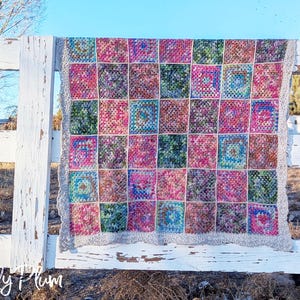 Puede incluir: Una manta de ganchillo con un borde blanco y un diseño de patchwork de cuadrados coloridos. La manta está colgada en una valla de madera blanca.