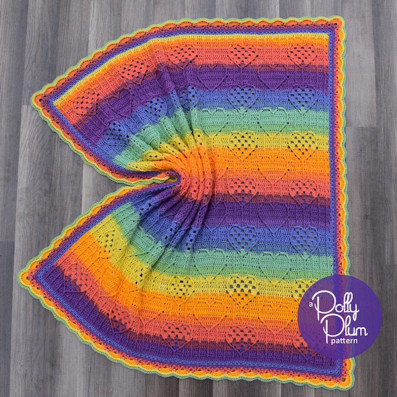 May include: A rainbow crochet blanket with a heart pattern and a scalloped edge. The blanket is folded in half and the text "a Dolly Plum pattern" is visible in the bottom right corner.