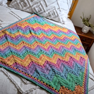 Può includere: Una coperta all'uncinetto colorata con un motivo a chevron in tonalità di rosa, viola, blu, verde, giallo e arancione. La coperta è appoggiata su un letto bianco con un cuscino bianco.