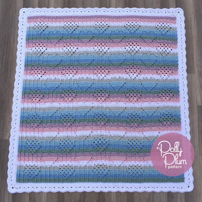May include: A crochet baby blanket with a white border and a heart pattern in shades of pink, blue, and green. The blanket has a scalloped edge and the text "a Polly Plum pattern" is in the lower right corner.