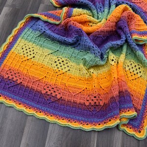 May include: A rainbow crochet blanket with a heart stitch pattern and a scalloped border. The blanket is made in shades of blue, green, yellow, orange, and purple.