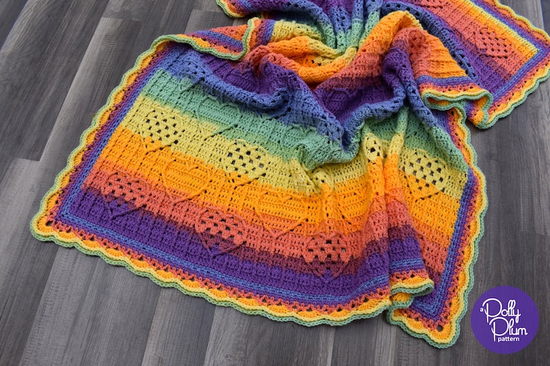 May include: A rainbow crochet blanket with a heart stitch pattern and a scalloped border. The blanket is made in shades of blue, green, yellow, orange, and purple.