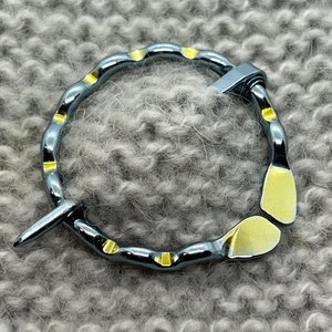 May include: A silver-colored shawl pin with yellow accents. The pin is circular with a pointed end and a hinged closure. The background is a gray knitted fabric.