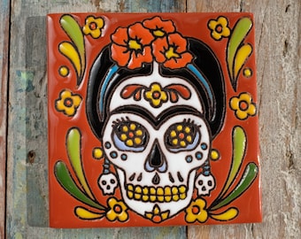 May include: A red ceramic tile with a colorful Day of the Dead sugar skull design. The skull is white with black outlines and features yellow, blue, and green details. The skull is surrounded by floral and leaf patterns in shades of red, orange, yellow, green, and blue.