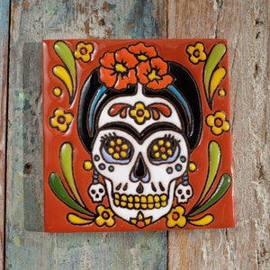 May include: A red ceramic tile featuring a colourful Day of the Dead sugar skull design. The skull is white with black outlines and has yellow, blue, and green details. The skull is surrounded by floral and leaf patterns in shades of red, orange, yellow, green, and blue.