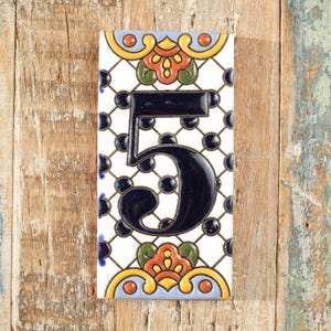 May include: Rectangular ceramic tile featuring the number 5 in dark blue. The tile has a white background with a black and white lattice pattern and colourful floral accents in orange, green, and yellow. The tile is set against a wooden background.
