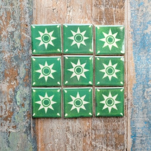 May include: Nine square green tiles arranged in a 3x3 grid. Each tile features a white starburst design with a central circle. The tiles are set against a weathered, multi-coloured wooden surface.