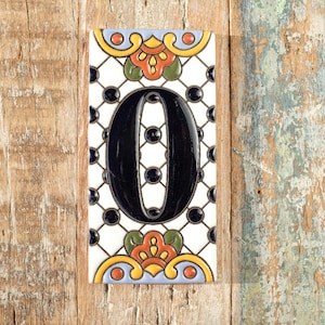 May include: Rectangular ceramic tile with the number "0" in black on a white background with a black dot grid. The tile has floral accents in orange, yellow, green, and blue. It is displayed on a wooden surface.