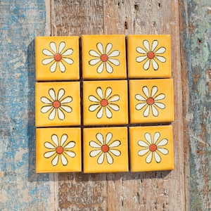 May include: Nine yellow ceramic tiles with white daisy designs and orange centres. The tiles are arranged in a 3x3 grid.