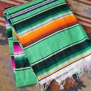 May include: A green, orange and black striped Mexican blanket with white fringe. The blanket is folded in half and has a pink and green striped section visible underneath.