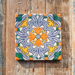 May include: Square ceramic tile with a floral design. The tile features a central orange flower surrounded by blue, yellow, and green floral patterns. The corners are accented with orange and green details. The tile is set against a rustic wooden background.