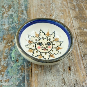 May include: A small, round ceramic bowl with a hand-painted sun design. The bowl has a white interior, a blue rim, and a brown outer edge. The sun has a face with pink cheeks and is surrounded by stylized rays.