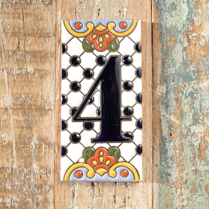 May include: Rectangular ceramic tile featuring the number 4 in black. The tile has a white background with a black dot pattern and decorative floral elements in orange, yellow, and green. The tile is displayed on a wooden surface.