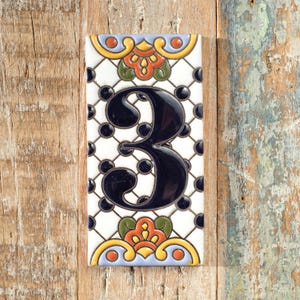 May include: A rectangular ceramic tile featuring the number 3 in navy blue. The tile has a white background with a black diamond pattern and navy blue dots. The top and bottom of the tile have colourful floral designs in orange, yellow, and green.