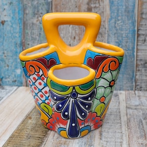 May include: A hand-painted ceramic utensil holder with a vibrant, colourful design. The holder features a yellow handle and rim, with a floral and geometric pattern in red, blue, green, and orange. It has three compartments and a decorative bow.