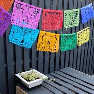 May include: A vibrant string of papel picado banners in various colours hangs against a dark grey wooden fence. The banners feature intricate cut-out designs in pink, red, blue, yellow, and green. A white planter box with greenery is positioned below.