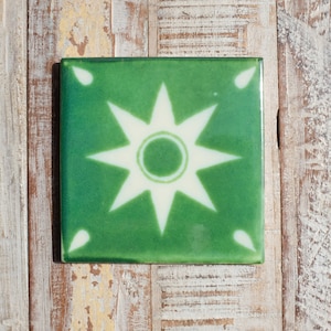 May include: Square green ceramic tile featuring a white starburst pattern. The tile has a glossy finish and is set against a weathered wooden surface. Decorative tile for home decor.