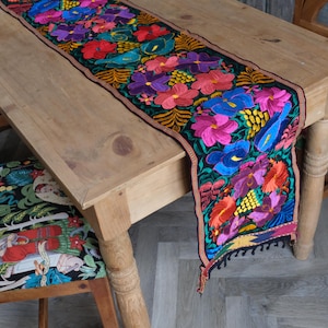 May include: A colourful floral embroidered table runner with a black background and fringe. The runner is on a wooden table.