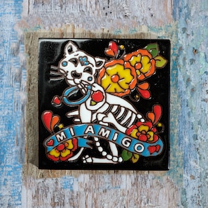 May include: A black and white ceramic tile featuring a colourful floral design and a skeleton cat. The cat is holding a blue ring and the tile reads "Mi Amigo".