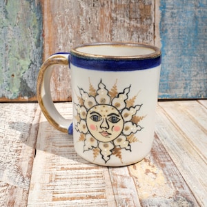May include: White ceramic mug featuring a hand-painted sun design. The sun has a smiling face with pink cheeks and a gold outline. The rim and handle are accented with blue and gold. The mug is on a weathered wooden surface.