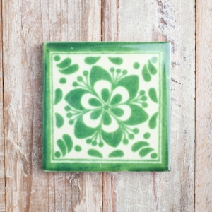 May include: Square ceramic tile featuring a green floral design on a cream background, framed by a green border. The tile is displayed on a light wood surface. The floral pattern is symmetrical and detailed.
