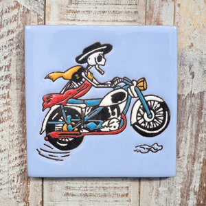 May include: A blue ceramic tile featuring a colourful illustration of a skeleton riding a motorbike. The skeleton is wearing a yellow and red scarf and a black hat. The motorbike is black and white with a blue seat.
